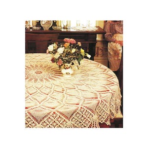 May include: A cream-colored, round crocheted tablecloth with a detailed floral and geometric pattern. The tablecloth is draped over a round table, with a vase of flowers in the center. The tablecloth has a scalloped edge.