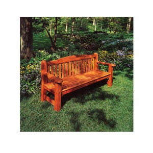 May include: A wooden park bench with a reddish-brown finish sits on a grassy lawn. The bench has a slatted back and armrests. The background features a garden with trees and flowers, creating a natural outdoor setting.