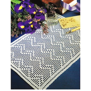 May include: A white, rectangular, crocheted doily with a detailed openwork pattern. The doily is displayed on a dark blue surface, alongside a bouquet of purple irises, a glass inkwell, and a vintage pen.