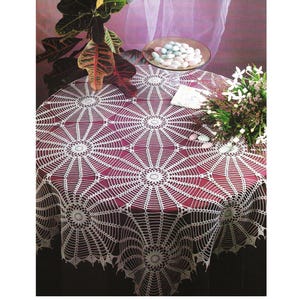 May include: A round table covered with a burgundy tablecloth featuring a white, intricate crochet spiderweb design. The table is set with a glass bowl of sugared almonds and a small bouquet of white flowers.