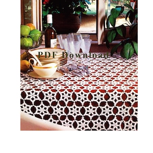 May include: A white crocheted tablecloth featuring a repeating star-like geometric pattern. The textile is draped over a table set with a stack of plates, a cream-coloured bowl with a gold rim, clear glassware, silverware, and a bottle of wine.