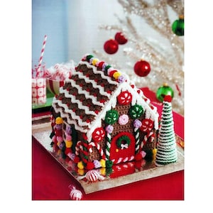 May include: A crocheted gingerbread house with a brown body, white roof accents, and colourful candy decorations. The house features a red door, a green wreath, and a small crocheted Christmas tree. The house sits on a silver tray.