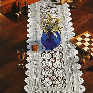 May include: White crocheted table runner with a delicate lace pattern. The runner is laid on a wooden table with a blue vase filled with white flowers and two glasses of wine.