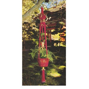 Puede incluir: Un colgador de plantas de macramé rojo con una cesta tejida que contiene plantas verdes. El colgador está suspendido de una rama de árbol, con una borla en la parte inferior. El fondo presenta un jardín japonés.