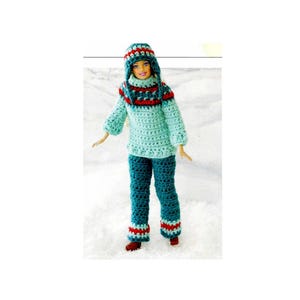 Puede incluir: Una muñeca con un conjunto de invierno de ganchillo hecho a mano. El conjunto incluye un suéter azul claro, pantalones a juego y un gorro, todos con detalles en rojo y azul oscuro. La muñeca está sobre una superficie blanca.
