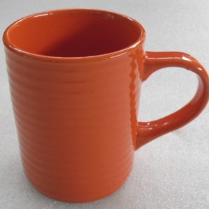 May include: A bright orange ceramic mug with a wide, rounded handle. The mug has a textured, ribbed design.