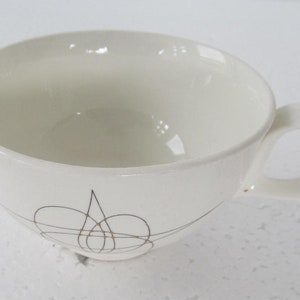 May include: White ceramic teacup with a gold and black abstract design. The cup has a rounded shape and a handle.