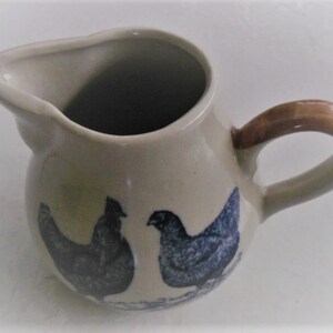 May include: A small, cream-colored ceramic creamer with a brown handle. It features a blue and white design of two chickens.