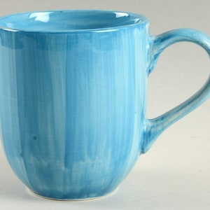 May include: A light blue ceramic mug with a rounded base and a curved handle. The mug has a glossy finish and a vertical brushstroke pattern in varying shades of blue. The mug is designed for hot or cold beverages.