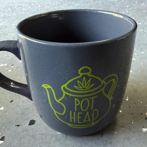 May include: A dark gray ceramic mug with a speckled finish and a handle on the left side. The front features a lime green graphic of a teapot with a leaf icon on the lid and the text POT HEAD printed in a bold, sans-serif font below the lid.