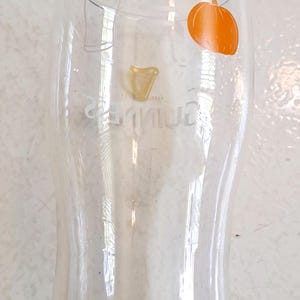 May include: A clear pint glass with a curved shape, featuring the Guinness logo and an orange pumpkin graphic. The glass has a wide top and a narrower base. The Guinness logo is in gold. The pumpkin is on the right side of the glass.
