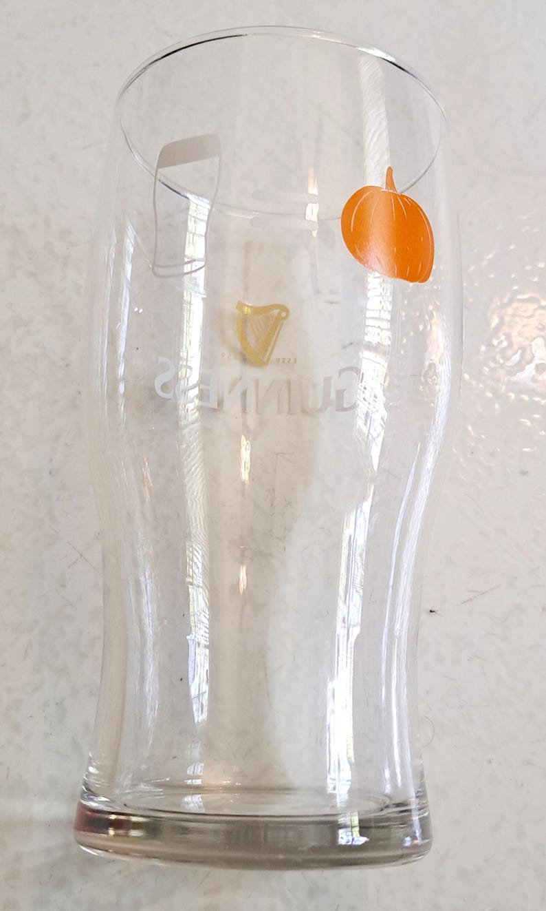 May include: A clear pint glass with a curved shape, featuring the Guinness logo and an orange pumpkin graphic. The glass has a wide top and a narrower base. The Guinness logo is in gold. The pumpkin is on the right side of the glass.