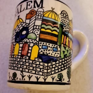 Puede incluir: Taza de cerámica blanca con una colorida ilustración de un paisaje urbano, con cúpulas, arcos y patrones geométricos. La palabra "LEM" está impresa en la parte superior. La taza tiene un asa blanca y un borde negro en la parte superior e inferior.