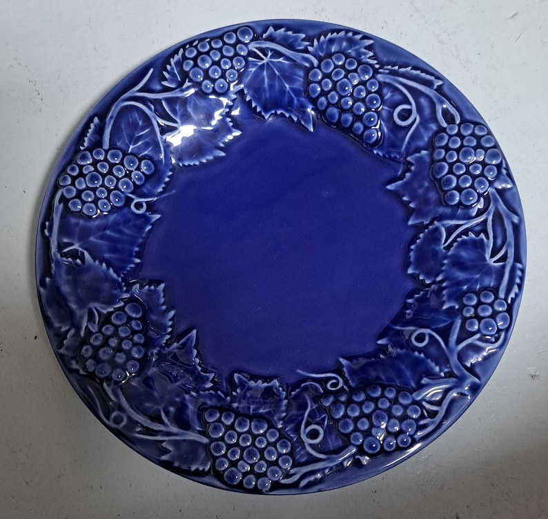 May include: A blue ceramic plate with a raised design of grapes and vines around the edge.
