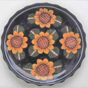 May include: A round, black ceramic plate with a scalloped edge. The plate is decorated with five orange sunflowers with green leaves. The center of each sunflower is a grid pattern in a darker orange color. The plate has a glossy finish.