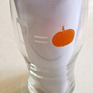 May include: Clear glass beer glass with a pumpkin graphic. The glass has a beer glass outline on the left, an equal sign in the middle, and an orange pumpkin on the right. The glass is empty and sitting on a white surface.