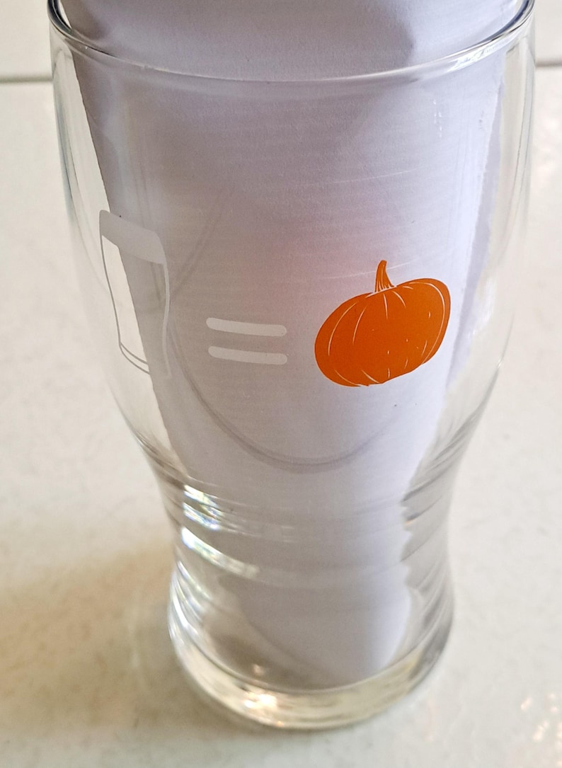 May include: Clear glass beer glass with a pumpkin graphic. The glass has a beer glass outline on the left, an equal sign in the middle, and an orange pumpkin on the right. The glass is empty and sitting on a white surface.