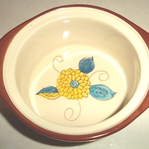 Puede incluir: Un pequeño cuenco de cerámica blanca con un borde marrón. El cuenco tiene un diseño floral pintado a mano en amarillo, azul y verde.