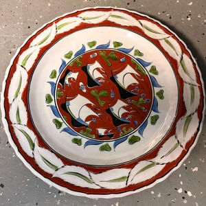 Peut inclure: Une assiette ronde en céramique avec un médaillon central rouge orné de voiliers stylisés blancs et noirs, entouré d'accents verts et bleus. Le large bord présente un motif répétitif de feuilles blanches sur un fond rouge.
