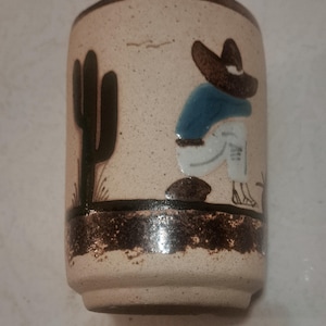 Pode incluir: Um copo de cerâmica com acabamento bege salpicado. O copo apresenta uma cena pintada de um cacto e uma figura usando um chapéu e camisa azul. A base e a borda são acentuadas com uma faixa marrom escura.