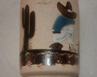 Puede incluir: Un vaso de cerámica con un acabado beige moteado. El vaso presenta una escena pintada de un cactus y una figura con sombrero y camisa azul. La base y el borde están acentuados con una banda marrón oscuro.