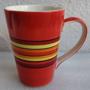 Può includere: Una tazza alta in ceramica rossa con manico bianco. La tazza ha un design a strisce in tonalità di giallo, arancione e rosso.
