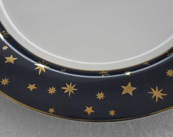 May include: A white ceramic plate with a dark blue border adorned with gold stars. The plate has a gold trim. The design is a celestial theme, suitable for serving food or as a decorative piece.