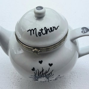 Può includere: Teiera in ceramica bianca con coperchio a cerniera. Il coperchio ha la scritta "Mother" in corsivo nero. La teiera ha un design in bianco e nero di una ragazza con cuori e una farfalla.
