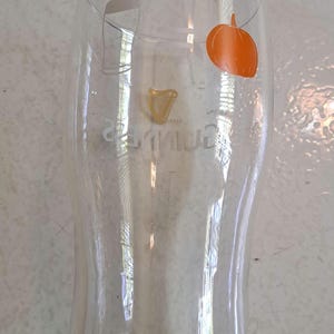May include: A clear Guinness pint glass with a curved shape. The Guinness logo is visible in gold, and an orange pumpkin sticker is on the glass. The glass is empty and sits on a light-colored surface.
