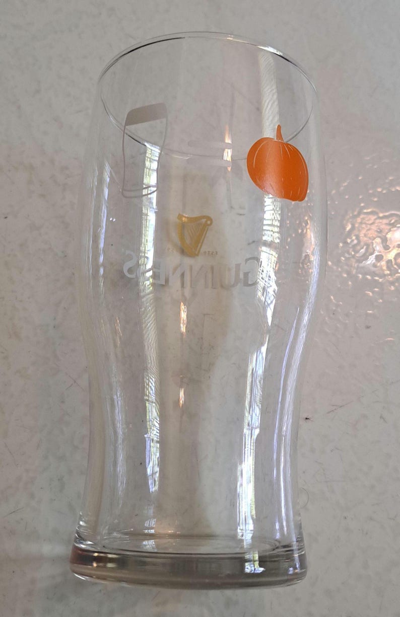 May include: A clear Guinness pint glass with a curved shape. The Guinness logo is visible in gold, and an orange pumpkin sticker is on the glass. The glass is empty and sits on a light-colored surface.