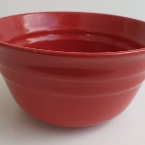 Puede incluir: Un cuenco de cerámica rojo brillante, de forma redondeada y con un borde ligeramente ensanchado. El cuenco tiene tres ranuras horizontales en el exterior. Adecuado para servir alimentos.