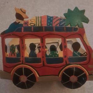 May include: A colorful wooden bus figurine with passengers and luggage on top. The bus is painted red with black wheels and yellow accents. Passengers are visible through the windows. A green star-shaped object is on the roof.