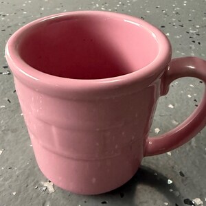 May include: A pink ceramic mug with a rounded rim and a curved handle. The mug has a cylindrical shape with horizontal ridges around the body. The interior of the mug is also pink.