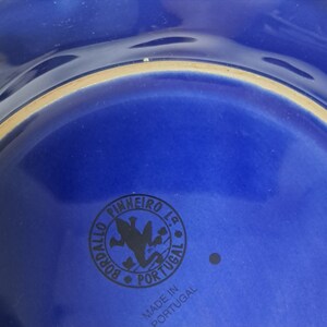 May include: Close-up of the bottom of a blue ceramic plate with a black logo that reads "Pinheiro Lda. Bordallo Portugal" and "Made in Portugal".