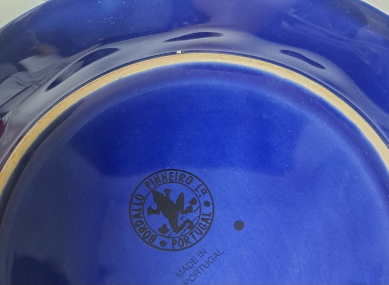 May include: Close-up of the bottom of a blue ceramic plate with a black logo that reads "Pinheiro Lda. Bordallo Portugal" and "Made in Portugal".