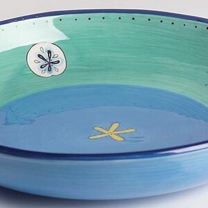 May include: A round, hand-painted ceramic bowl with a coastal theme. The bowl features a light blue exterior, a two-tone interior of teal and blue, and is decorated with starfish and sand dollar designs. The rim is outlined with a dark blue border.