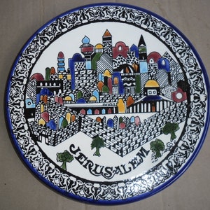 May include: A white ceramic plate with a blue and white border. The center of the plate features a colorful illustration of the city of Jerusalem with the word "Jerusalem" written in black below the illustration.