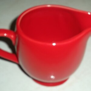 May include: A bright red ceramic creamer with a rounded body and a handle.