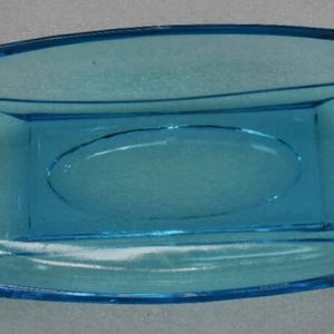 May include: A rectangular, light blue glass dish with rounded ends. The dish has a slightly raised oval in the center and curved details on the sides. The overall design is simple and elegant, suitable for serving or display.