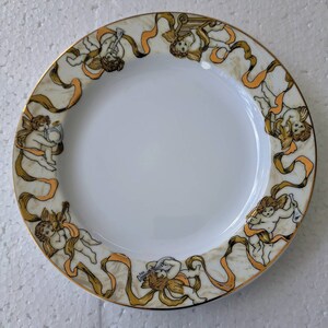 Pode incluir: Prato de porcelana branca com borda dourada e uma borda decorativa de querubins tocando instrumentos musicais. Os querubins são cercados por fitas douradas sobre um fundo creme.