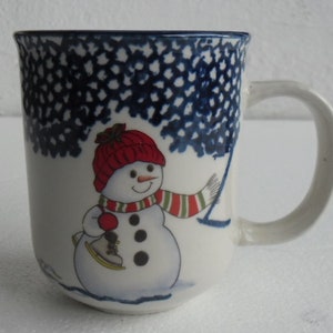 May include: White ceramic mug with a blue and white snowflake design. The mug features a snowman wearing a red hat and a green and white striped scarf. The snowman is holding a red pom-pom and a blue ice skate.