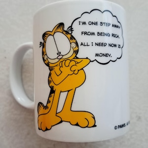 May include: White ceramic mug featuring Garfield, the orange cartoon cat, with the text "I'm one step away from being rich. All I need now is money." The mug has a white handle and is a fun, novelty item.