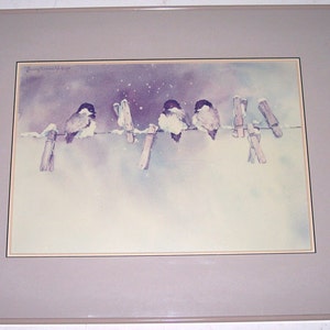 May include: A watercolor painting of five birds perched on a clothesline with clothespins. The birds are a light purple color with dark eyes and beaks. The background is a light purple with white snow falling. The painting is framed in a light brown frame.