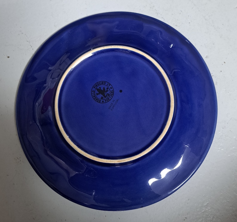 May include: Blue ceramic plate with a white rim and a small black stamp on the bottom that reads "Pinheiro 18, Portugal, Made in Portugal."