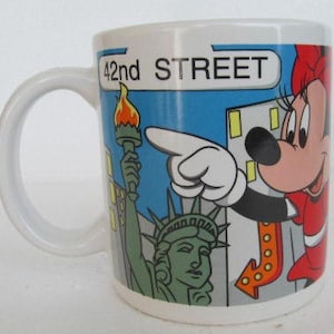 May include: White ceramic mug with a colorful illustration of Mickey Mouse pointing to a sign that says "42nd STREET". The illustration also includes the Statue of Liberty and a cityscape.