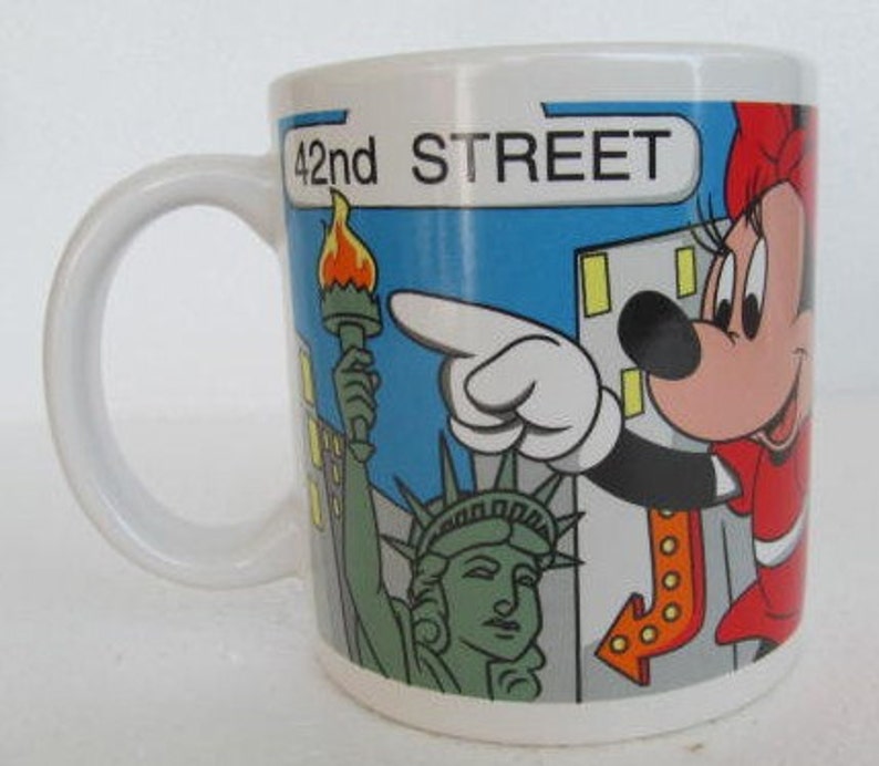 May include: White ceramic mug with a colorful illustration of Mickey Mouse pointing to a sign that says "42nd STREET". The illustration also includes the Statue of Liberty and a cityscape.