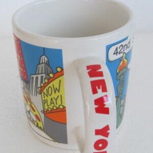 May include: White ceramic mug with a colorful illustration of New York City landmarks, including the Empire State Building, the Statue of Liberty, and a yellow taxi. The handle of the mug says "NEW YORK" in bold, white letters.