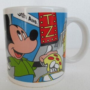 May include: White ceramic mug with a colorful illustration of Mickey Mouse in a green shirt, holding a slice of pizza. The background features a blue sky, yellow taxi, and a red sign with the text "T.Z."