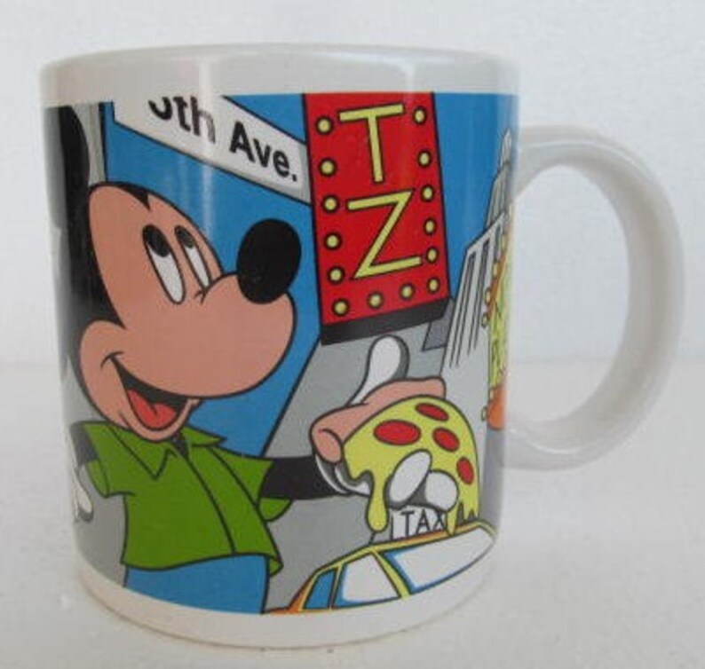May include: White ceramic mug with a colorful illustration of Mickey Mouse in a green shirt, holding a slice of pizza. The background features a blue sky, yellow taxi, and a red sign with the text "T.Z."