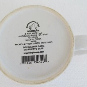 May include: White ceramic mug with a black and white image of Mickey Mouse and the text "Everyone loves to get applause" on the front. The back of the mug has the text "Applause, LLC Woodland Hills, CA 91367 &copy; Disney Made in China 34320 Mickey & Friends New York Mug Dishwasher Safe. Microwave Safe. www.applause.com" and a barcode.